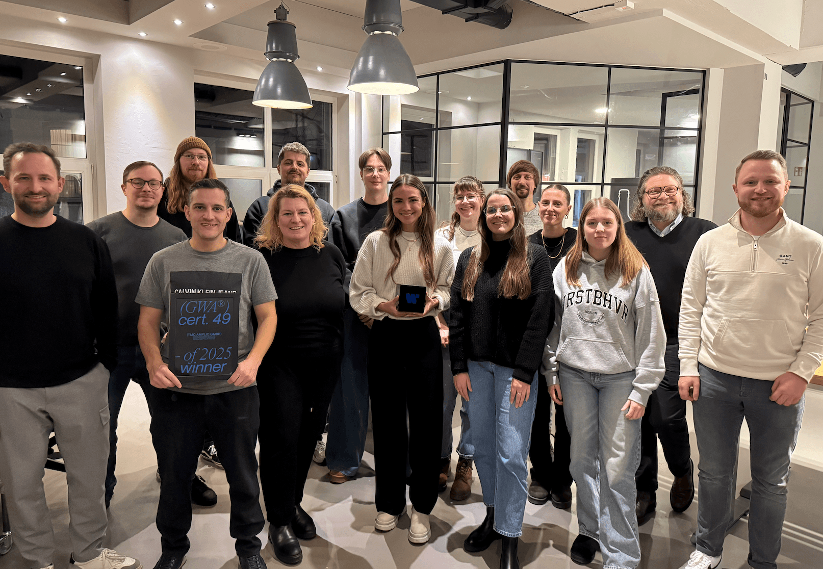 Gruppenfoto in einem Büro: Mehrere Personen stehen nebeneinander in einem hellen Raum mit Glaswänden; einige lächeln in die Kamera, im Hintergrund sind Arbeitsbereiche und Deckenleuchten zu sehen.