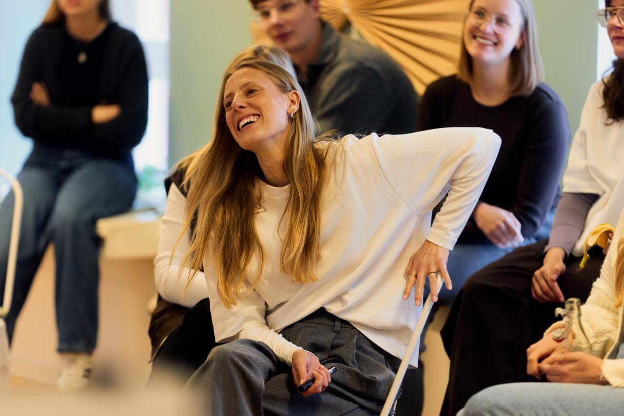 Teilnehmende sitzen in einem Workshop; eine Person im Vordergrund lacht und lehnt sich entspannt zurück.