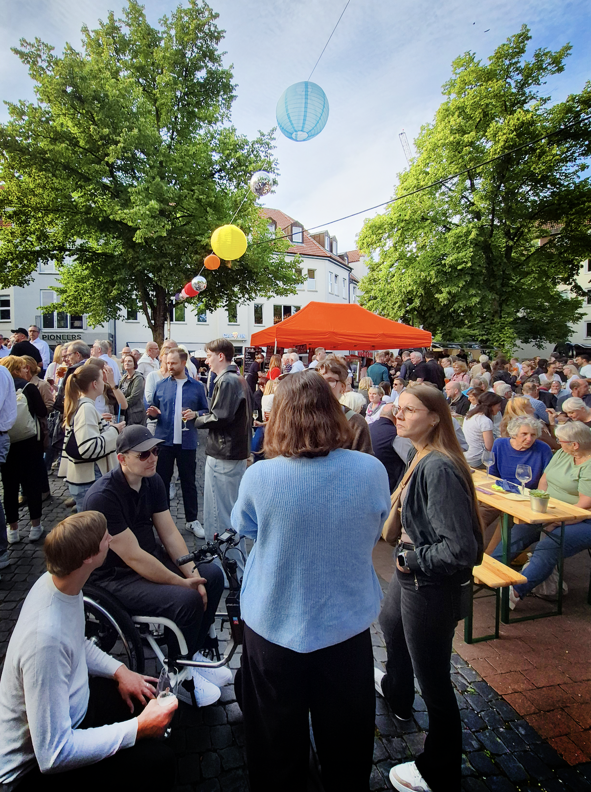 Menschenmenge bei einer Outdoor-Veranstaltung in der Stadt: Viele Personen sitzen und stehen auf einem Platz zwischen Bäumen, im Mittelpunkt ein roter Sonnenschirm.
