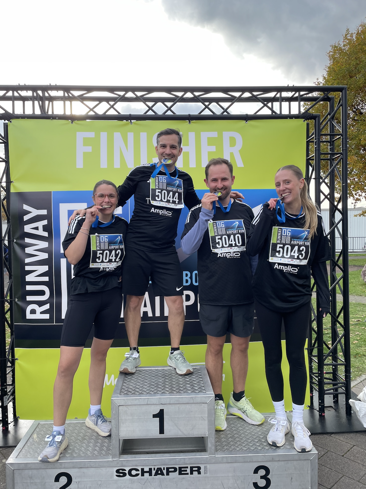 Vier Läuferinnen und Läufer posieren nach einem Lauf nebeneinander vor einer grünen FINISHER-Wand, tragen Medaillen und halten Urkunden; im Vordergrund steht ein Siegerpodest.
