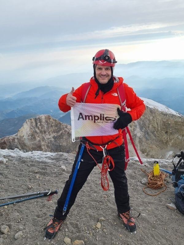 Person in roter Outdoor-Kleidung steht am Gipfelkreuz auf einem felsigen Berggipfel, hält ein Schild mit der Aufschrift „Amplio“; im Hintergrund sind Berge und Himmel zu sehen.