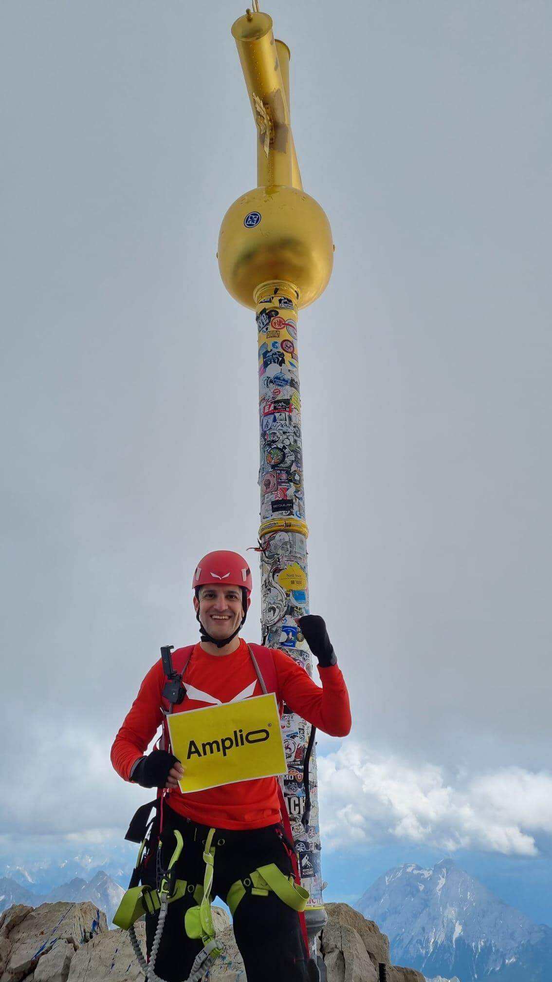 Bergsteiger in roter Jacke und Helm steht auf der Zugspitze vor dem goldenen Gipfelkreuz, hält ein Schild „Amplio“; im Hintergrund ein weiter Blick auf Berge unter blauem Himmel.
