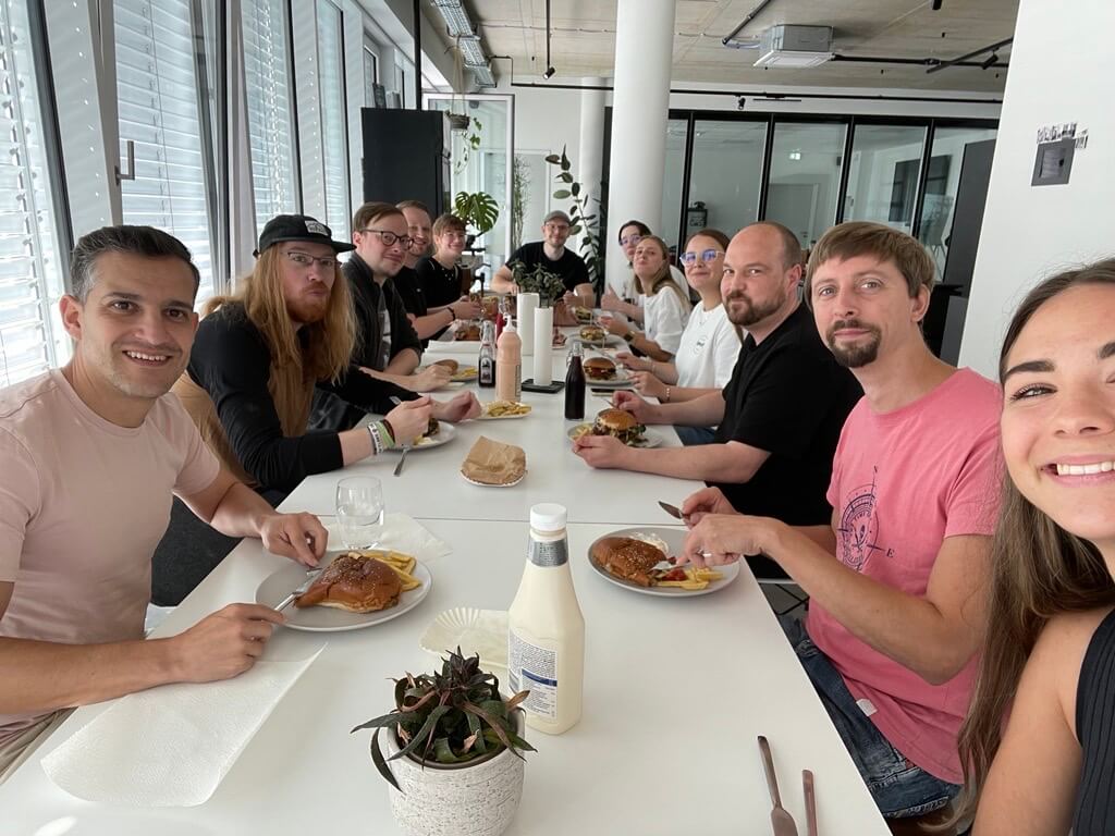 Mehrere Personen sitzen an einem langen Tisch beim gemeinsamen Essen in einem hellen Raum; Teller mit Speisen stehen vor ihnen, alle schauen in die Kamera.