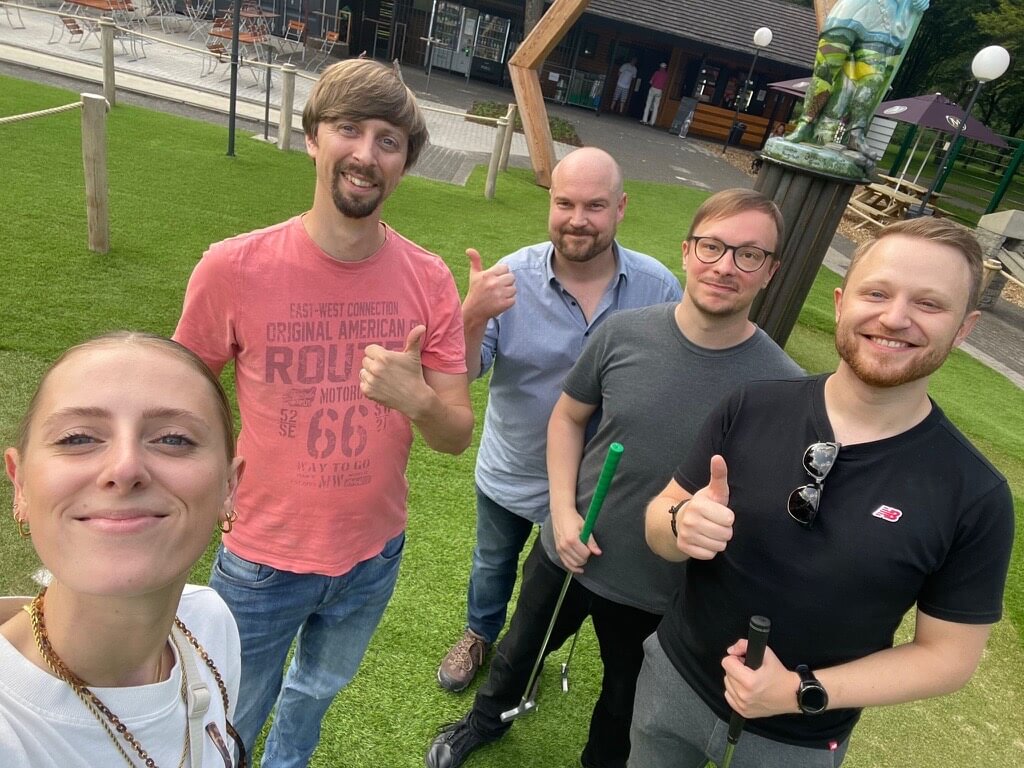 Fünf Personen stehen draußen auf einer Wiese und machen ein Gruppen-Selfie; einige zeigen den Daumen nach oben, im Hintergrund sind Gebäude und ein Sonnenschirm zu sehen.