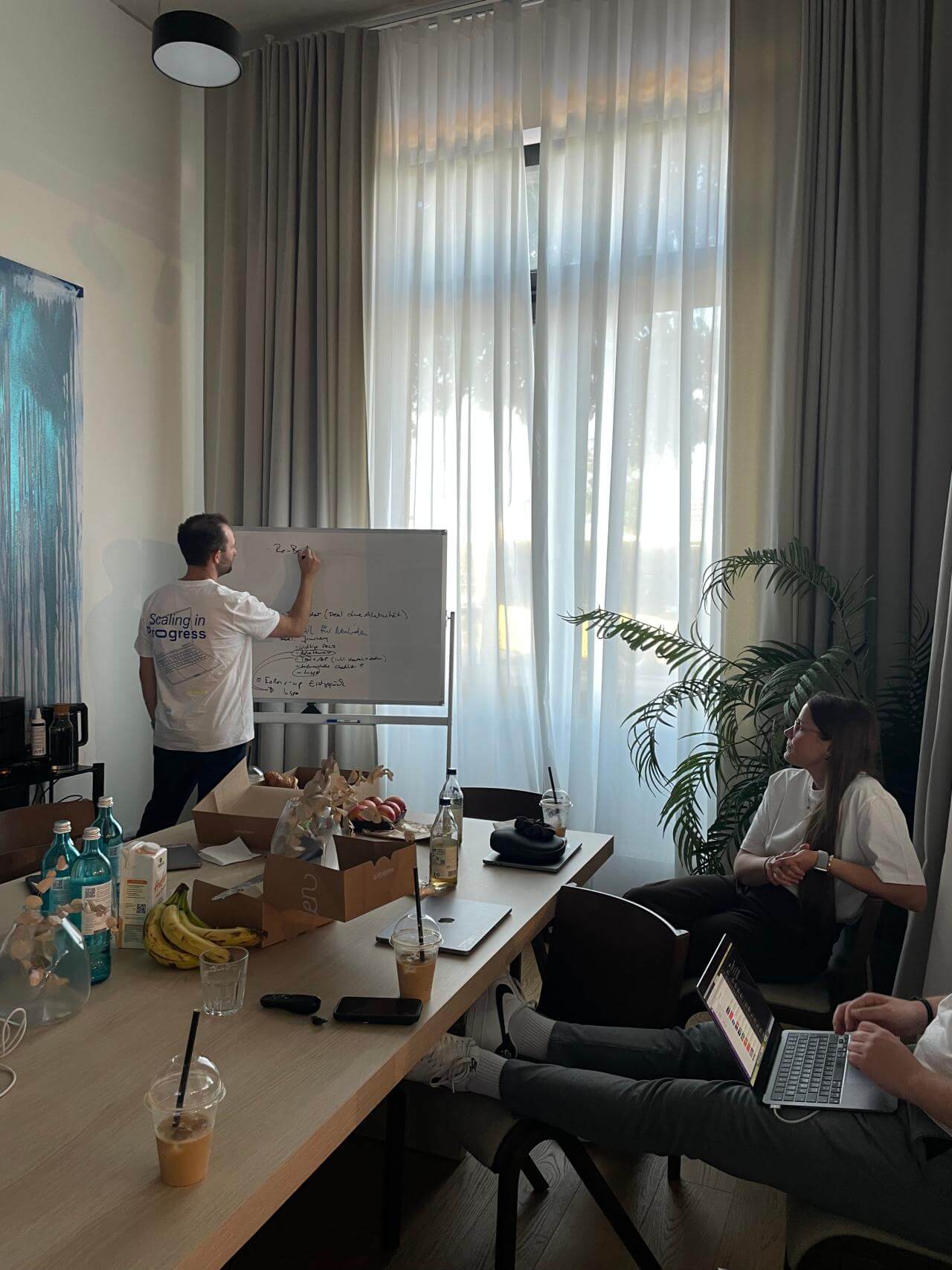 Workshop im Büro: Eine Person steht am Flipchart und schreibt, zwei weitere sitzen am Tisch und hören zu; auf dem Tisch stehen Getränke und Snacks, großes Fenster mit Vorhängen im Hintergrund.