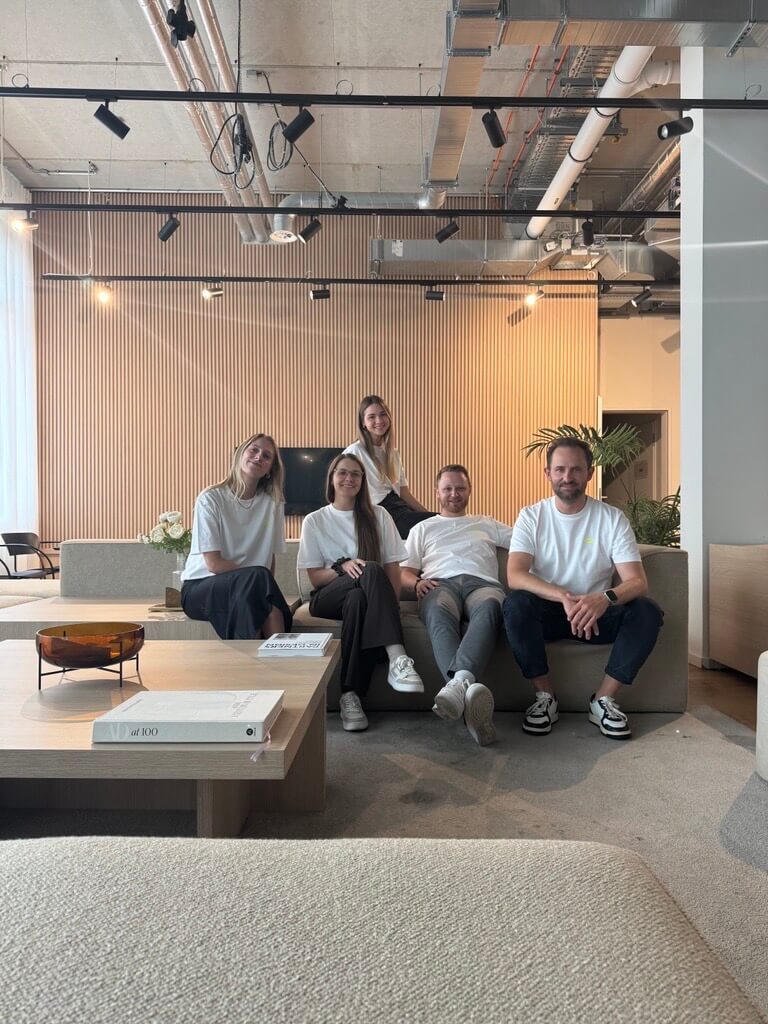 Fünf Personen sitzen nebeneinander auf einem Sofa in einem modernen, hellen Hotel-Lobby mit sichtbarer Decke und Lampen; im Vordergrund stehen niedrige Tische.