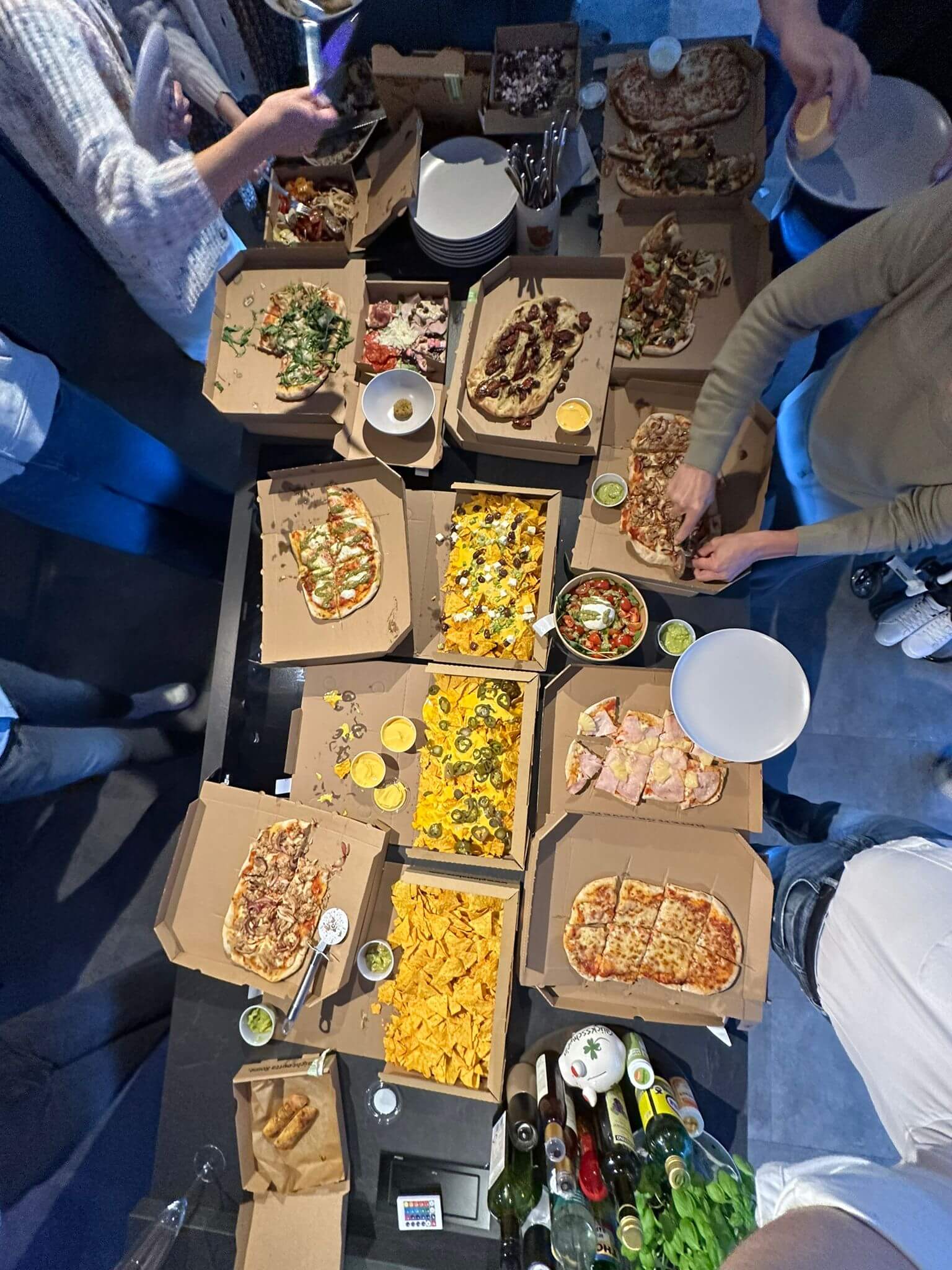 Blick von oben auf einen Tisch mit mehreren offenen Pizzakartons, Salat und Getränken; mehrere Personen stehen darum herum und greifen nach den Speisen.