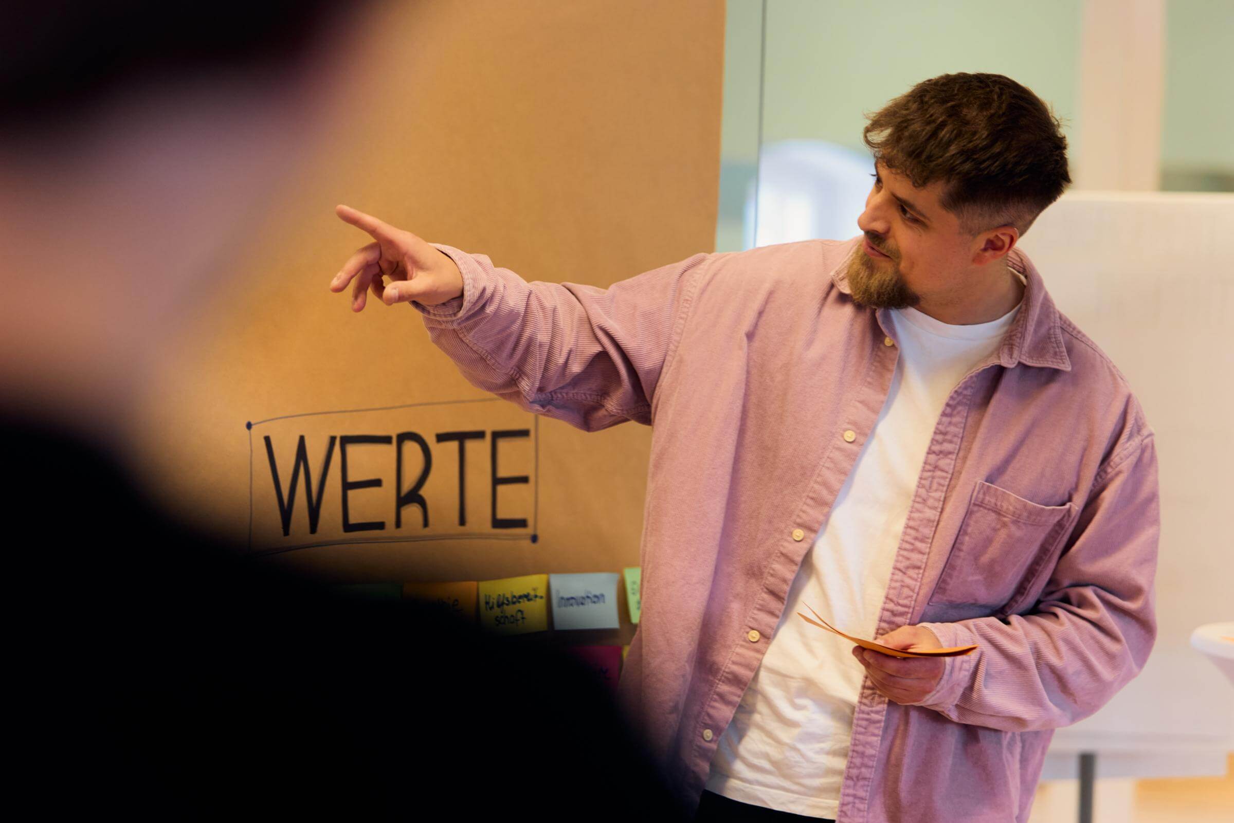 Mann in rosa Hemd zeigt während einer Präsentation auf ein Board mit der Überschrift „WERTE“, unscharfe Person im Vordergrund, warmes Licht im Raum.