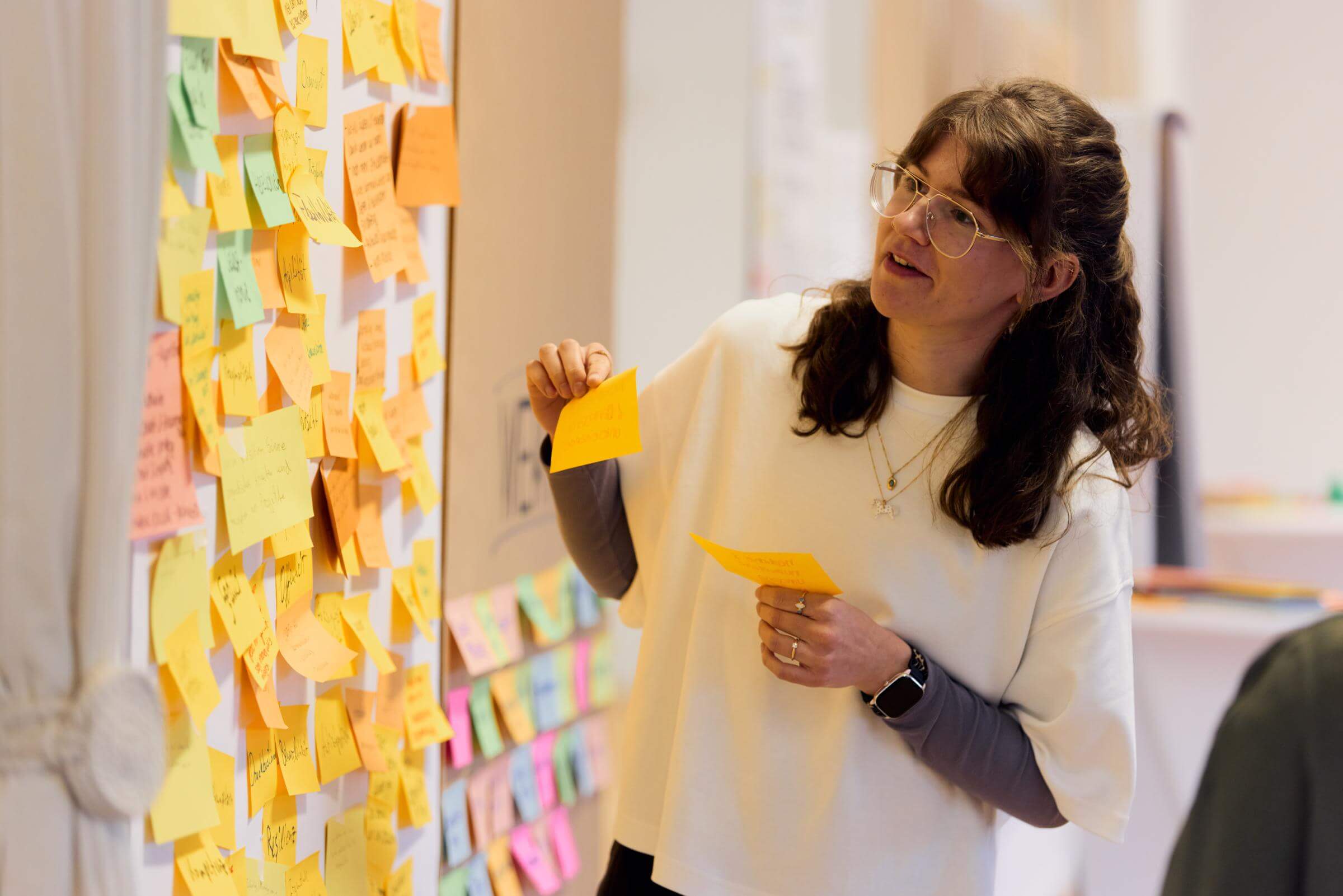 Frau mit Brille hängt Notizzettel an eine Wand voller bunter Post-its, hält einen Stapel Zettel in der Hand; Workshop-Szene in hellem Raum.