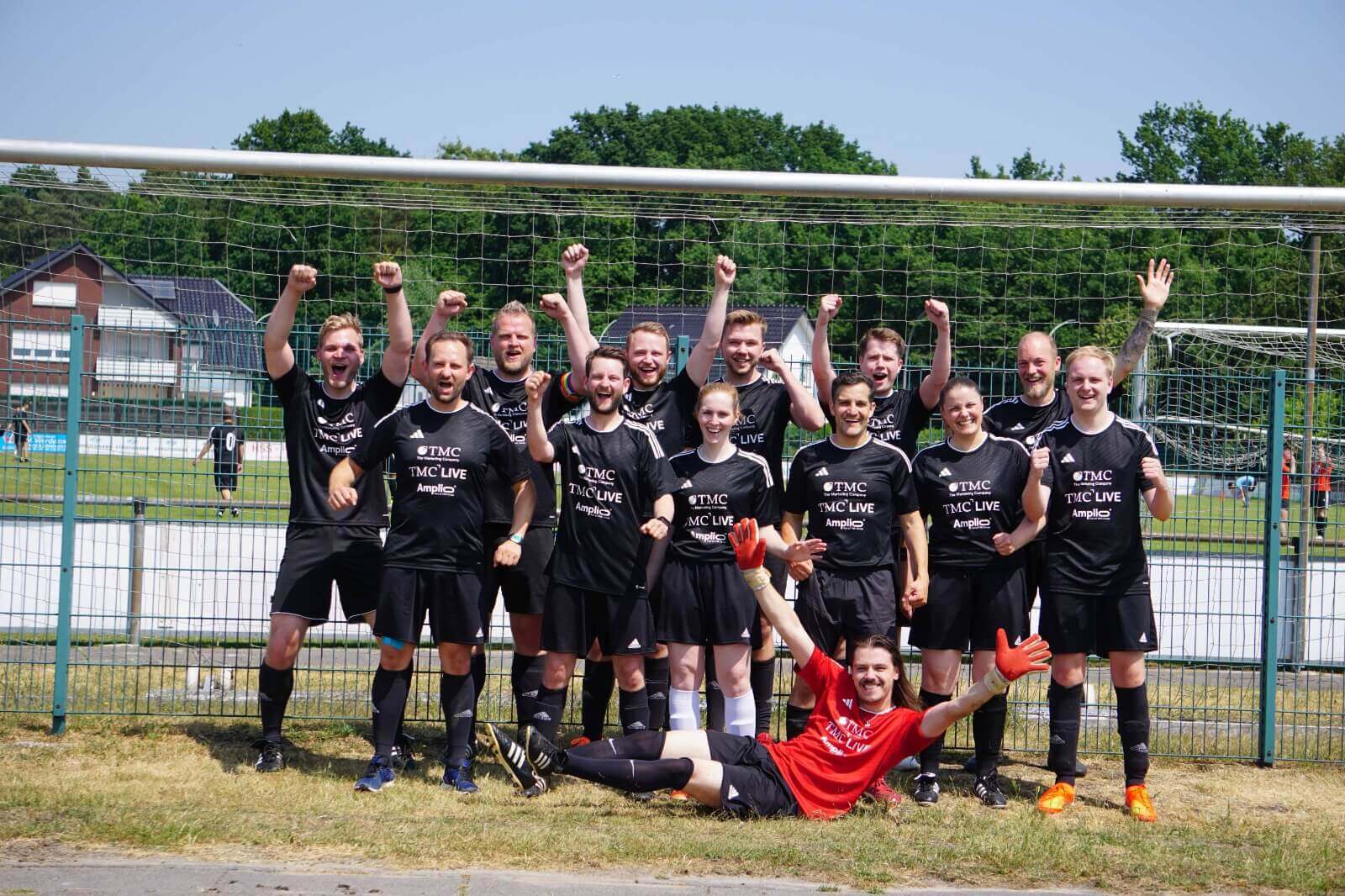 Gruppenfoto einer Fußballmannschaft auf dem Rasen: Team in schwarzen Trikots steht und kniet in Reihen, eine Person in Rot liegt vorne; im Hintergrund Zaun und Sportplatz.