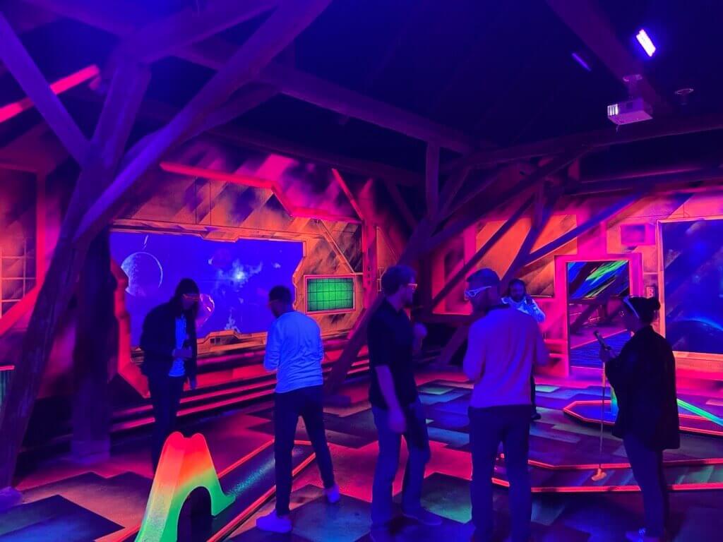 Gruppe von Personen beim Schwarzlicht-Minigolf in einer Indoor-Location mit UV-Beleuchtung; Wände und Boden leuchten in bunten Neonfarben, mehrere Personen stehen an der Bahn.