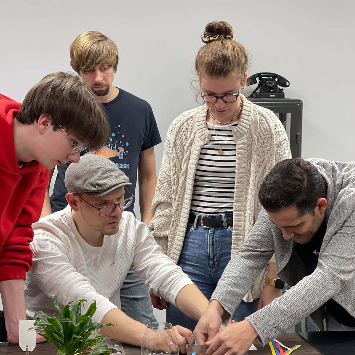 Mehrere Personen stehen an einem Tisch und lösen gemeinsam Rätsel bei einem Exit-Game; sie beugen sich über Materialien auf dem Tisch, im Vordergrund steht eine kleine Pflanze.